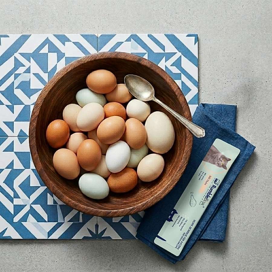 Handcrafted wooden bowl filled with assorted colored eggs on blue patterned mat, accompanied by a silver spoon and napkin.