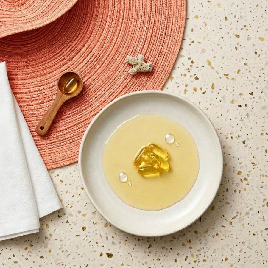 Golden omega-3 fish oil capsules in a white bowl with honey-like oil, set on a speckled countertop and coral placemat.